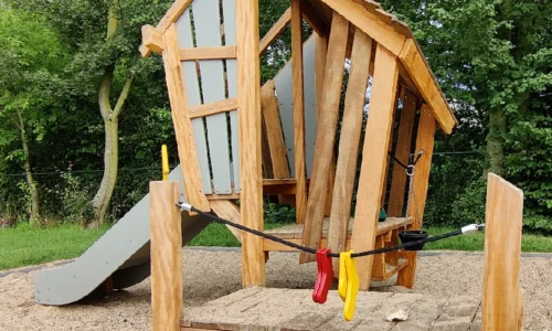 Levendige foto van een speelhut van het merk Kloek, aangeboden door LuduQ; een natuurlijk houten speelhuisje met kleurrijke details zoals een oven, waslijn en glijbaan, ontworpen voor kinderen tot 6 jaar.