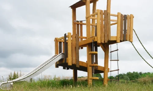Levendige foto van het speeltoestel Vos van het merk Kloek, aangeboden door LuduQ; een houten klim- en speeltoestel met natuurlijke lijnen, glijbaan en klimgedeelte, waarop kinderen actief klimmen en spelen in een groene buitenomgeving.
